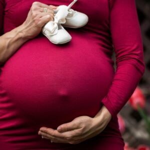 Pregnant woman holding baby shoes on belly in garden setting.